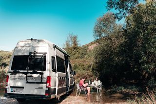 Ein Grand California 600 Van steht beim Überwintern in Portugal an einer Lichtung. Ein Mann und eine Frau sitzen im Bach daneben an einem Campingtisch und essen.