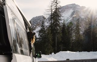 Eine Frau schaut beim Wintercamping aus einem geparkten Grand California Van auf eine verschneite Bergkulisse.