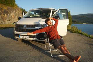 Island Rundreise 10 Tage: Junger Mann streckt sich auf seinem Campingstuhl vor einem Off Camper am Ufer eines Sees