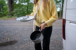 Eine junge Frau in gelbem Pulli trägt dreckige Teller, Besteck und eine Pfanne zum Abwasch - mit sauberem Wassertank im Wohnmobil.