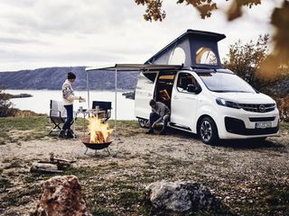 Ein Opel Crosscamp an einem See geparkt. Zwei Männer bereiten neben einem Lagerfeuer ihr Abendessen vor.