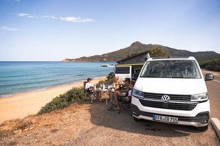 Zwei Opel Crosscamp Vans stehen geparkt neben einer Küstenstraße. Eine Freundesgruppe sitzt davor an einem Campingtisch und scheint zu frühstücken.