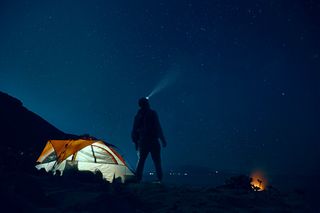 Eine Person steht in der Nacht vor einem Zelt und leuchtet mit der Stirnlampe in den Nachhimmel. 