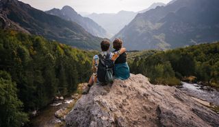 Zwei Freunde sitzen beim Wandern bei ihrer Albanien Rundreise Arm in Arm auf einem Felsen und betrachten die Aussicht auf die Berge.