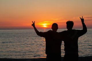 Ein schwules Paar auf ihrer Reise mit dem Camper: vor dem Sonnenuntergang am Meer zeigen sie ein Peace-Zeichen. 