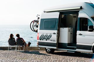 Eine Frau und ein Mann sitzen in Campingstühlen neben einem VW Grand California Van, der an einer Küstenstraße geparkt ist.
