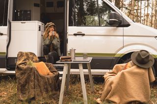 Ein Grand California Van steht geparkt in einem herbstlichen Wald. Eine Frau sitzt in der geöffneten Schiebetür mit einem warmen Getränk, vor dem Van sind ein Campingtisch und -stühle aufgebaut.