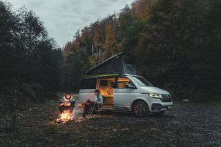 Ein VW Ocean Van steht in einem herbstlichen Wald, neben ihm stehen ein Campingtisch und -stühle, am Tisch sitzt eine Freundesgruppe.