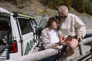 Zwei junge Frauen lehnen an einem Zaun und umarmen sich, im Hintergrund ein weißer Off Camper