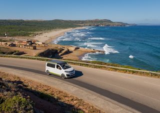 Ein VW California Van fährt eine Küstenstraße entlang. Im Hintergrund erstreckt sich das blaue Meer.