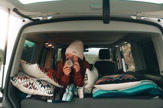 The back of a CamperBoys van is opened and there are lots of pillows. A woman in winter clothing inside the vehicle is drinking something hot out of a CamperBoys mug.