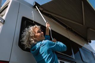 Camping Wissen: Eine Frau mit grauen Locken fährt die Markise ihres Campers aus und lacht dabei.