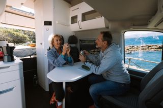 Strom im Wohnmobil ist das A und O einer Camperreise: Ein Ehepaar sitzt im Wohnraum des Campers am Meer, unterhält sich und trinkt Kaffee.