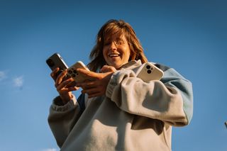 Eine junge Frau hält drei Handys gleichzeitig in den Händen und lacht vor dem strahlend blauen Himmel im Hintergrund.