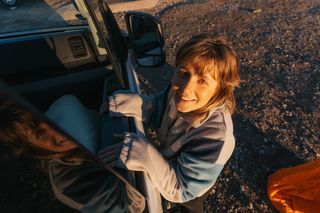 Eine junge Frau beim Campen für Anfänger steht neben der Beifahrertür des Campers und schaut lachend in die Kamera nach oben, während ihr die Sonne ins Gesicht scheint.